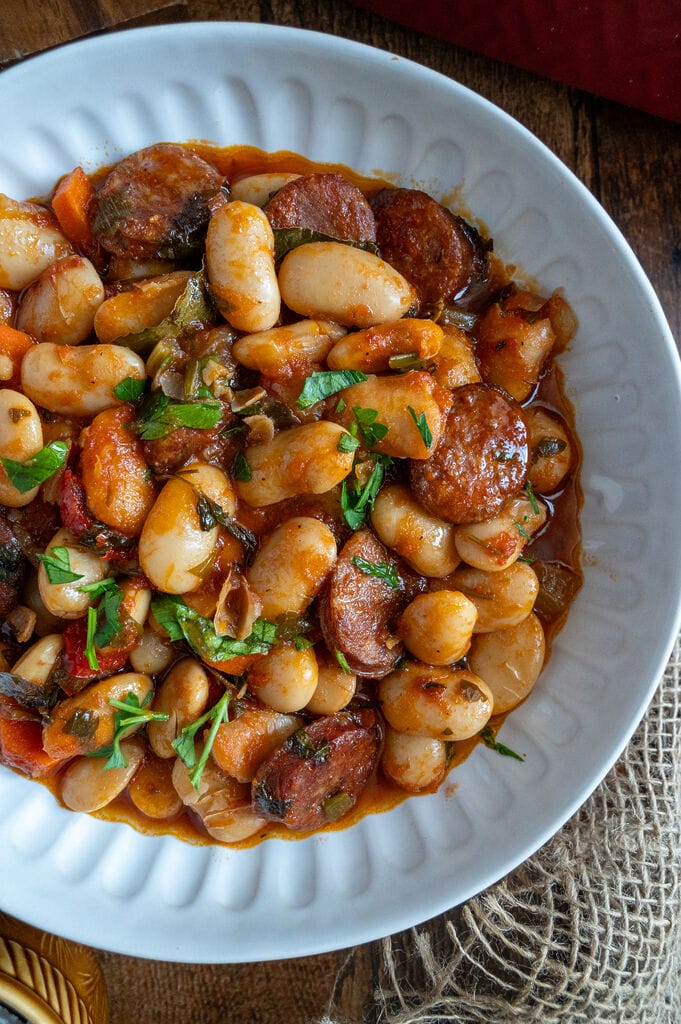 Greek Baked Gigantes with Sausage (Loukaniko) in a white bowl, surrounded by bread, and olives, and feta.