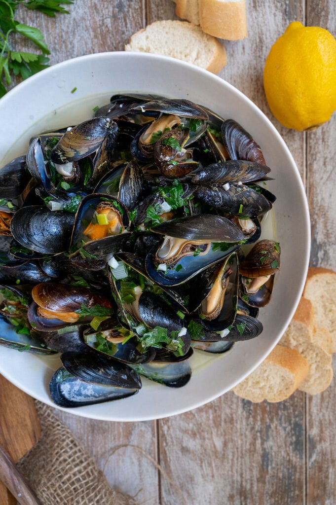 Steamed Mussels in White Wine Sauce in a white bowl surrounded by bread.