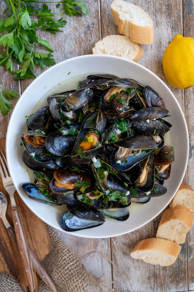 Steamed Mussels in White Wine Sauce in a white bowl surrounded by bread.