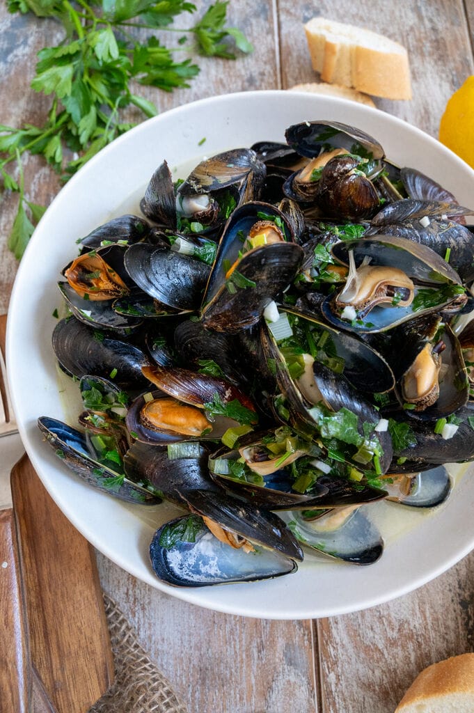 Steamed Mussels in White Wine Sauce in a white bowl surrounded by bread.