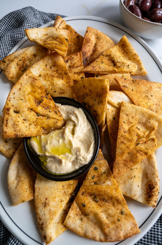 Pita chips on a white platter along with a small bowl of humus, surrounded by olives.
