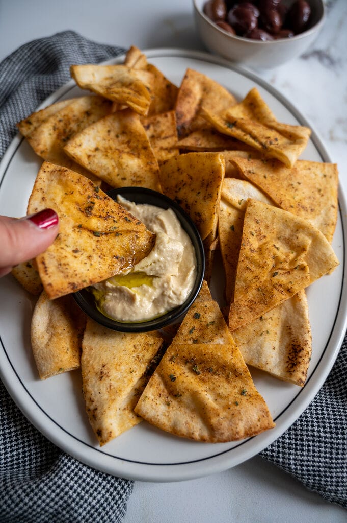 Pita chips on a white platter along with a small bowl of humus, surrounded by olives.