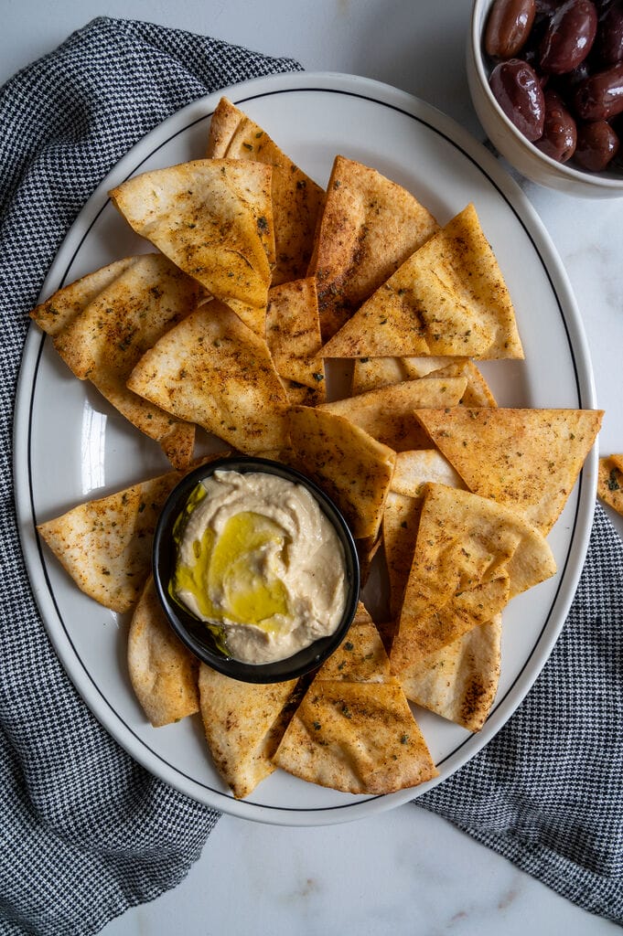 Pita chips on a white platter along with a small bowl of humus, surrounded by olives.
