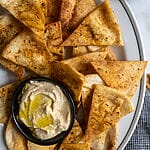 Pita chips on a white platter along with a small bowl of humus, surrounded by olives.