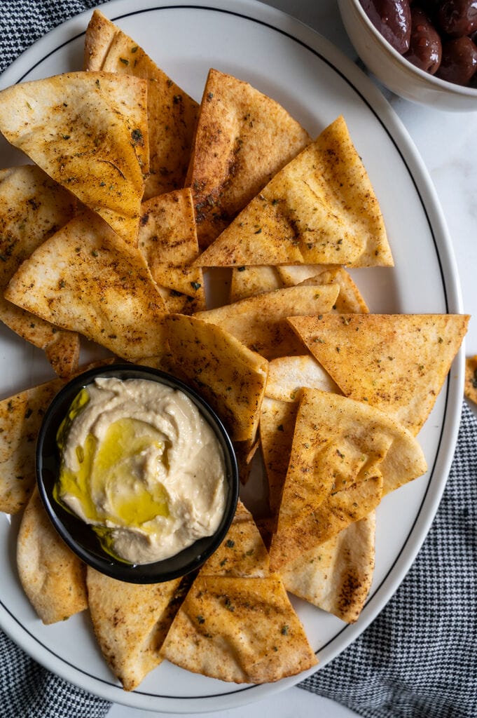 Pita chips on a white platter along with a small bowl of humus, surrounded by olives.