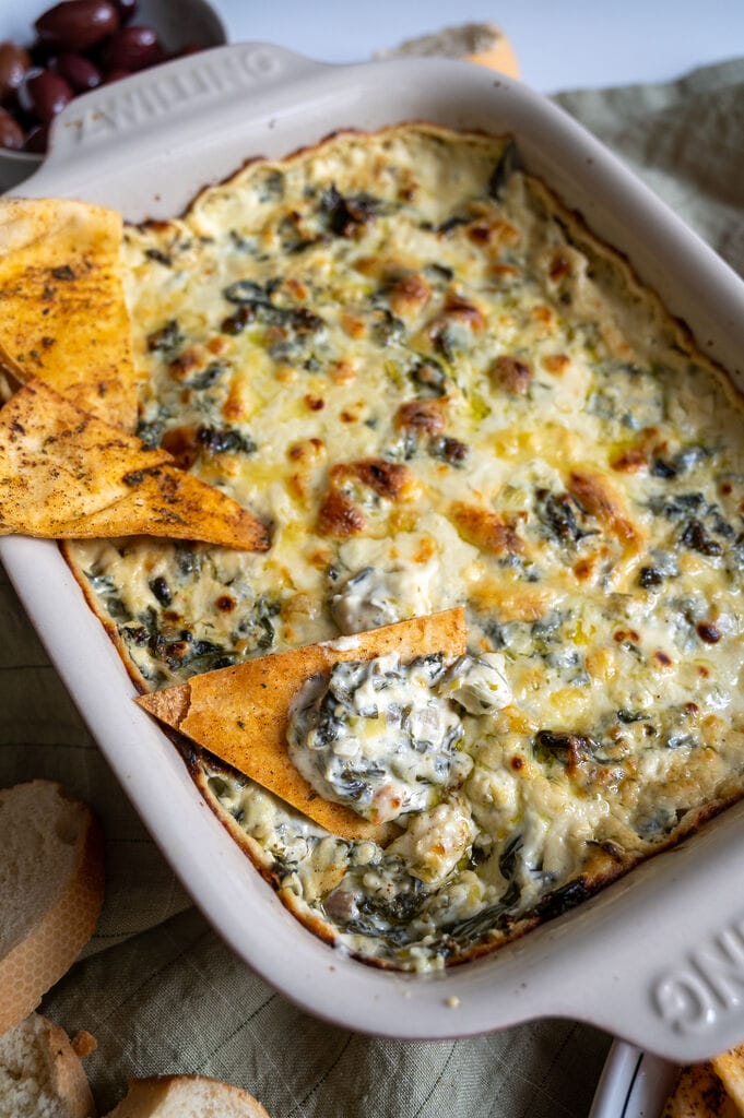 Baked spanakopita dip in a white casserole dish, with 3 baked pita chips on it.