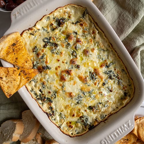 Baked spanakopita dip in a white casserole dish, with 2 baked pita chips on it.