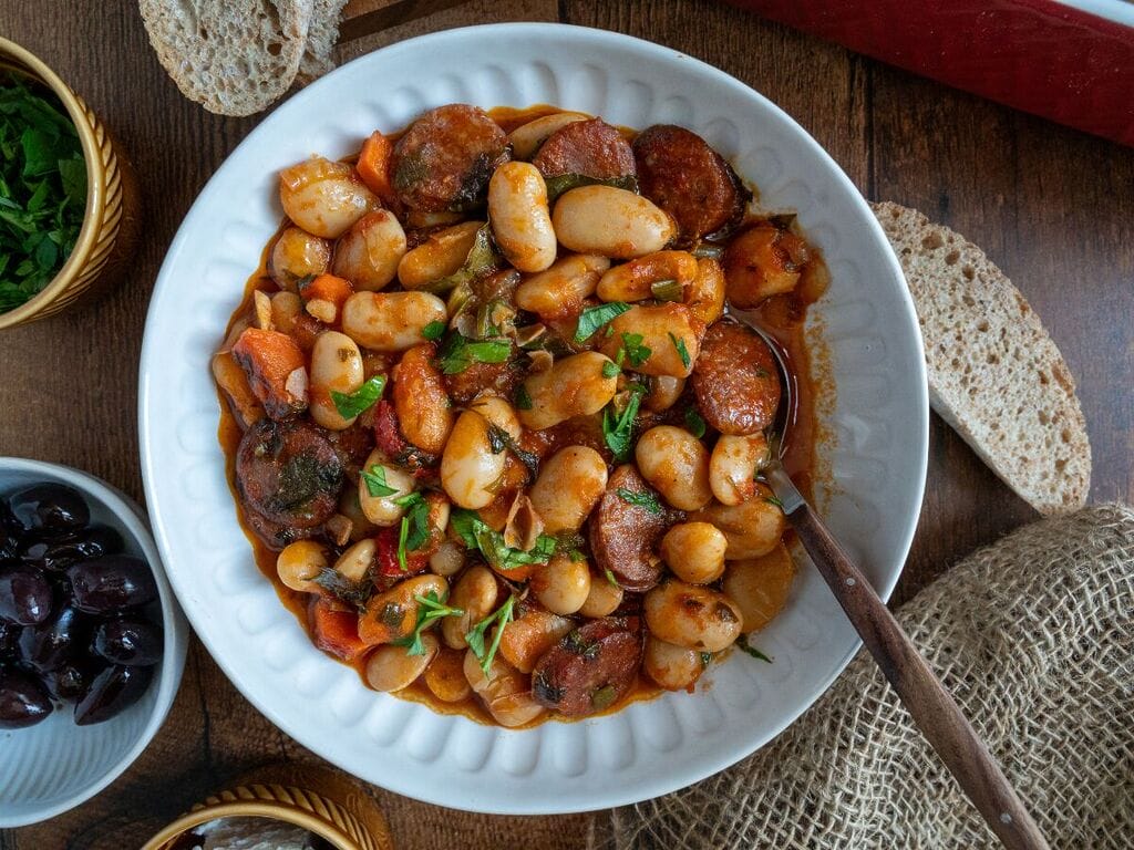 Greek Baked Gigantes with Sausage (Loukaniko) in a white bowl, surrounded by bread, and olives, and feta.