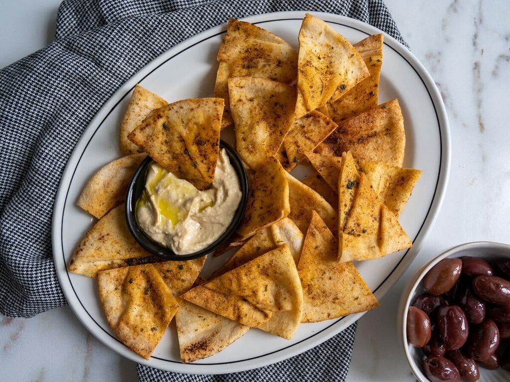 Pita chips on a white platter along with a small bowl of humus, surrounded by olives.