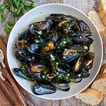 Steamed Mussels in White Wine Sauce in a white bowl surrounded by bread.