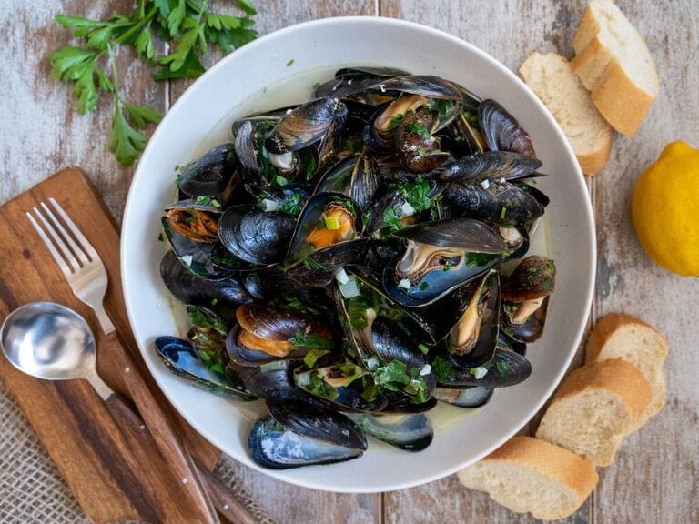 Steamed Mussels in White Wine Sauce in a white bowl surrounded by bread.