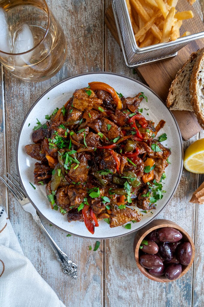 Greek Pork Tigania with Peppers on a white plate, surrounded by olives, fries, bread, and extra lemon.