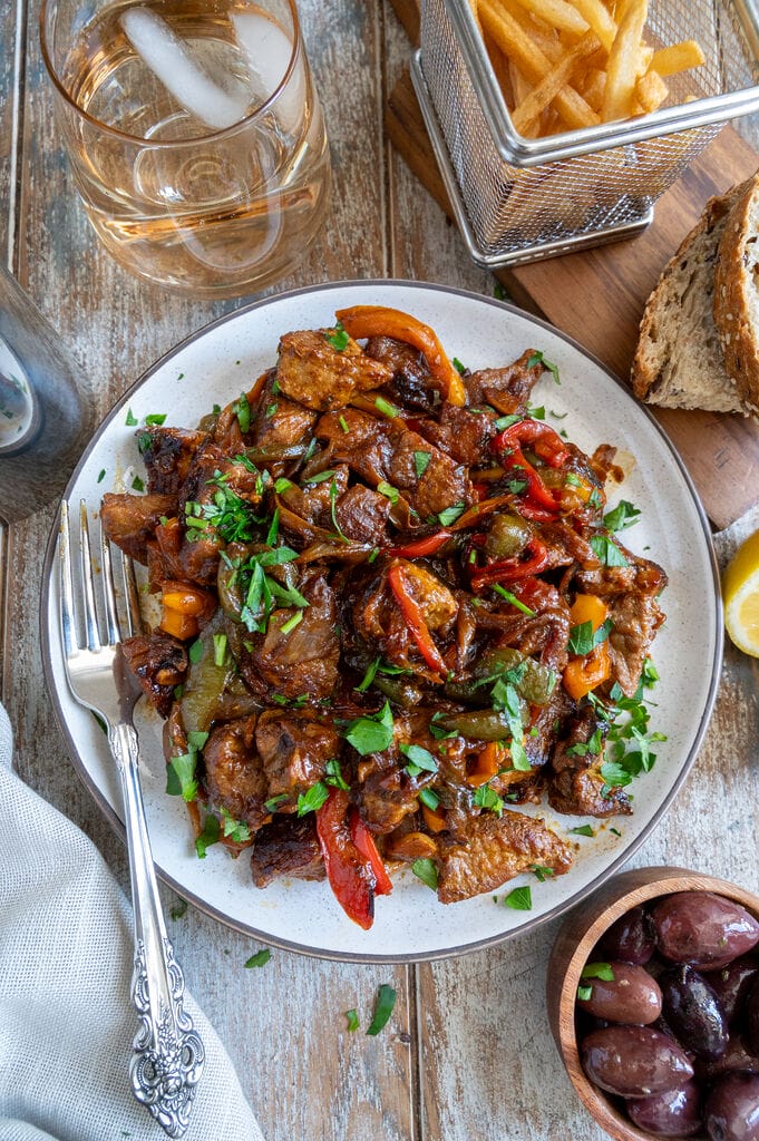 Greek Pork Tigania with Peppers on a white plate, surrounded by olives, fries, bread, and extra lemon.