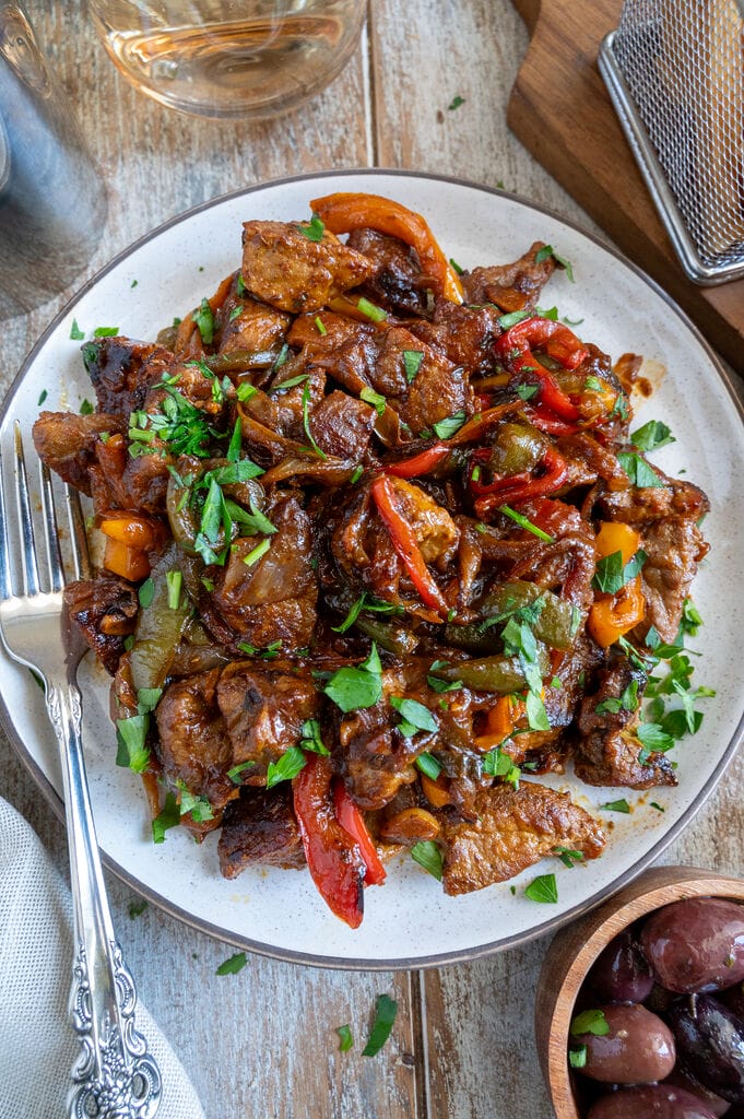 Greek Pork Tigania with Peppers on a white plate, surrounded by olives, fries, bread, and extra lemon.
