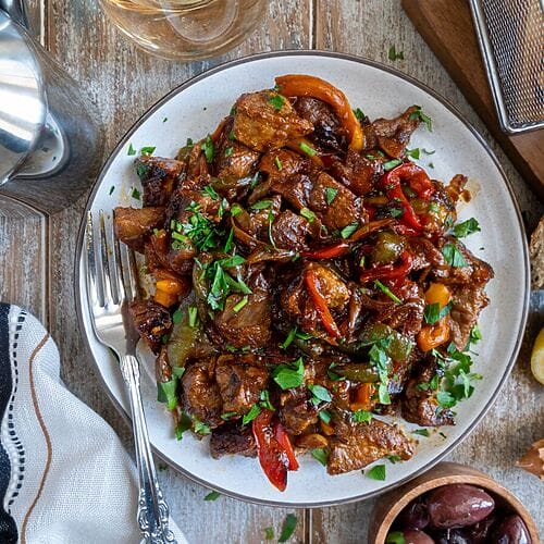 Greek Pork Tigania with Peppers on a white plate, surrounded by olives, fries, bread, and extra lemon.