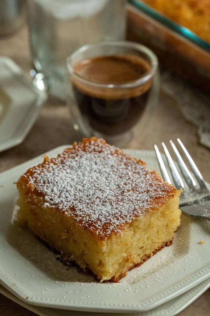 A piece of lemonopita cake on a white plate, and topped with a sprinkle of icing sugar, with a Greek coffee next to it.