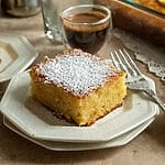 A piece of lemonopita cake on a white plate, and topped with a sprinkle of icing sugar, with a Greek coffee next to it.