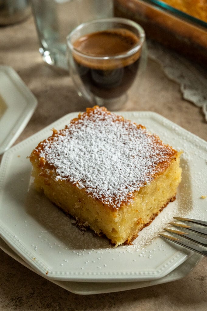 A piece of lemonopita cake on a white plate, and topped with a sprinkle of icing sugar, with a Greek coffee next to it.