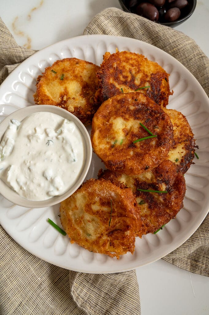 Fried potato fritters on a white plate with a small bowl of tzatziki and some olives on the side.