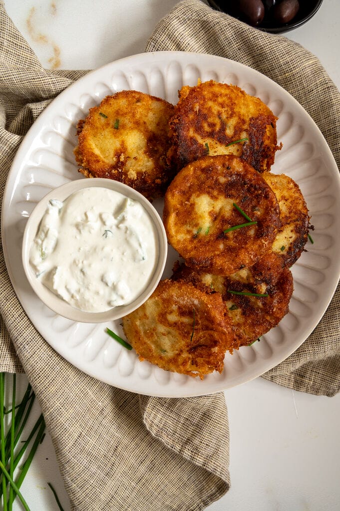 Fried potato fritters on a white plate with a small bowl of tzatziki and some olives on the side.