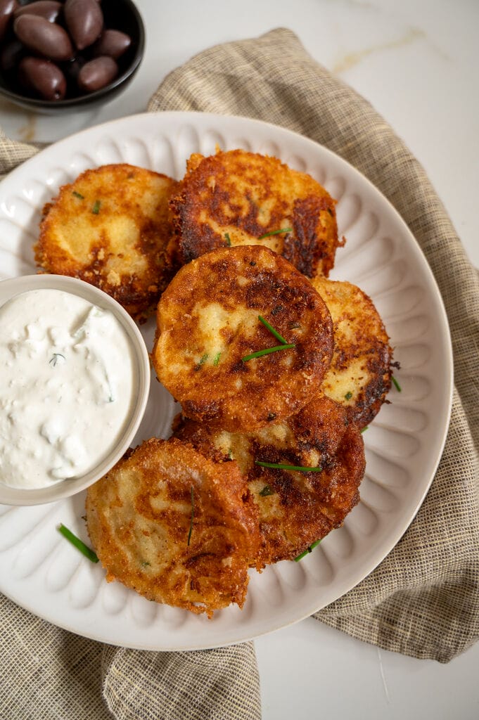 Fried potato fritters on a white plate with a small bowl of tzatziki and some olives on the side.