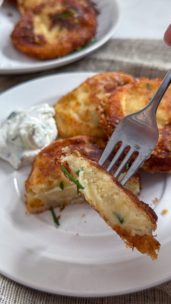 Fried potato fritters on a white plate with a small bowl of tzatziki and some olives on the side. Showing the inside of the fritter.