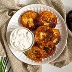 Fried potato fritters on a white plate with a small bowl of tzatziki and some olives on the side.