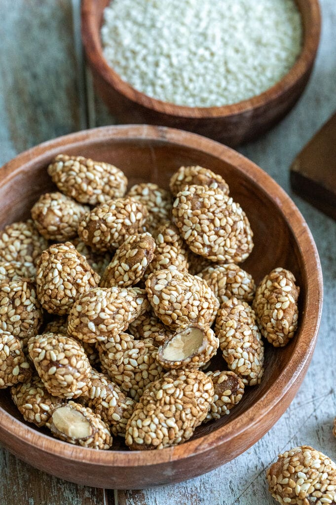 Greek sesame coated caramelized almonds, in a brown wooden bowl surrounded by bowls of sugar, almonds and sesame seeds.
