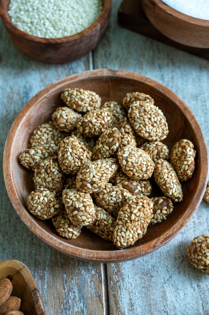 Greek sesame coated caramelized almonds, in a brown wooden bowl surrounded by bowls of sugar, almonds and sesame seeds.