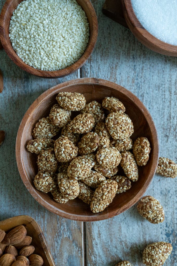 Greek sesame coated caramelized almonds, in a brown wooden bowl surrounded by bowls of sugar, almonds and sesame seeds.