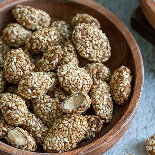 Greek sesame coated caramelized almonds, in a brown wooden bowl surrounded by bowls of sugar, almonds and sesame seeds.