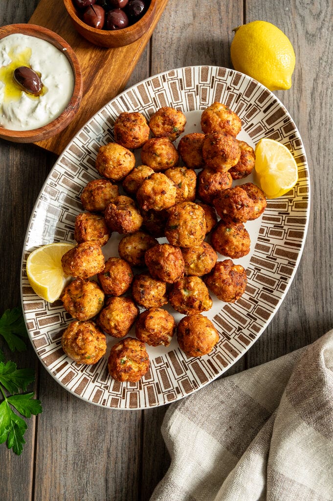 Greek chicken meatballs with feta on a white and brown platter, surrounded by lemons, tzatziki and olives.