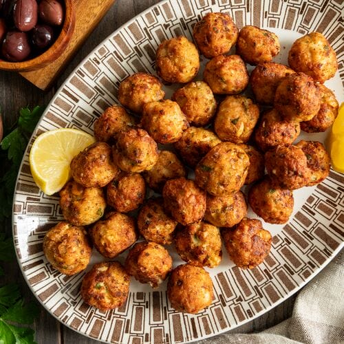 Greek chicken meatballs with feta on a white and brown platter, surrounded by lemons, tzatziki and olives.