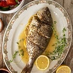 The baked sea bream on a white platter, surrounded by juices, lemon slices and fresh thyme.