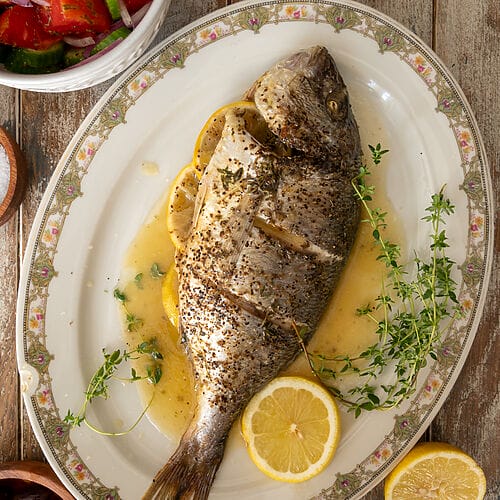 The baked sea bream on a white platter, surrounded by juices, lemon slices and fresh thyme.