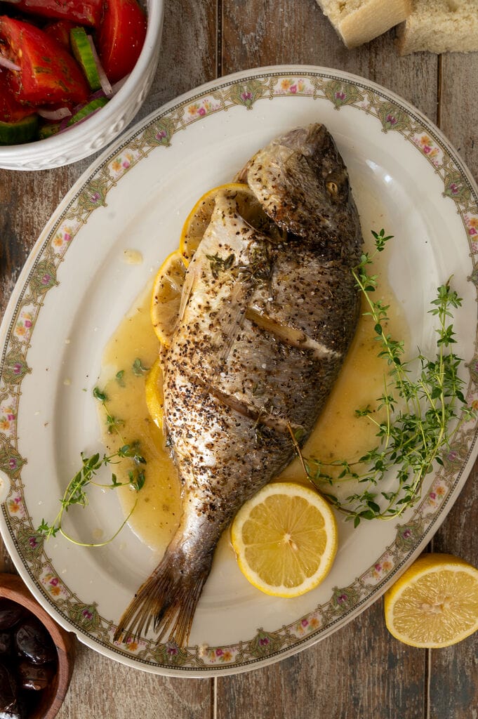 The baked sea bream on a white platter, surrounded by juices, lemon slices and fresh thyme.